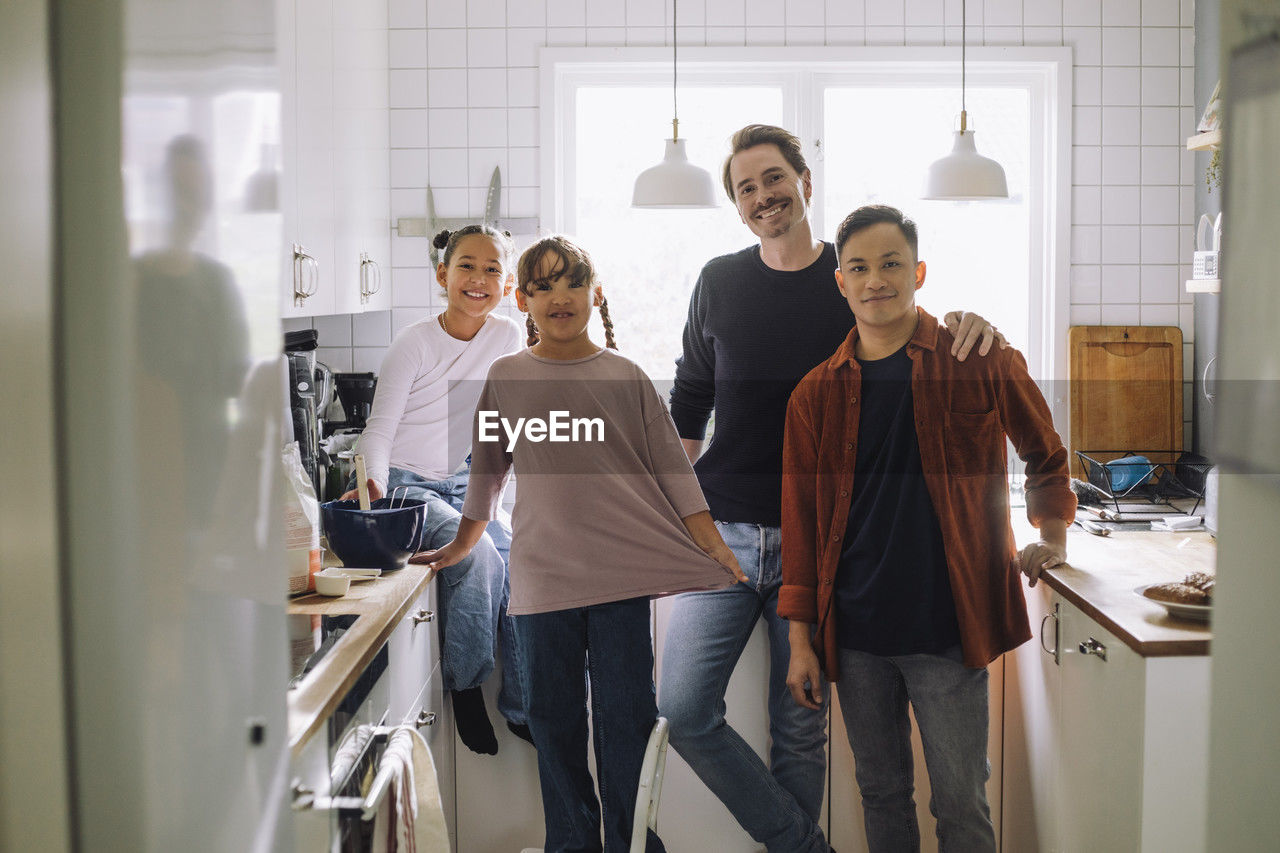 Portrait of happy gay men standing with daughters in kitchen at home