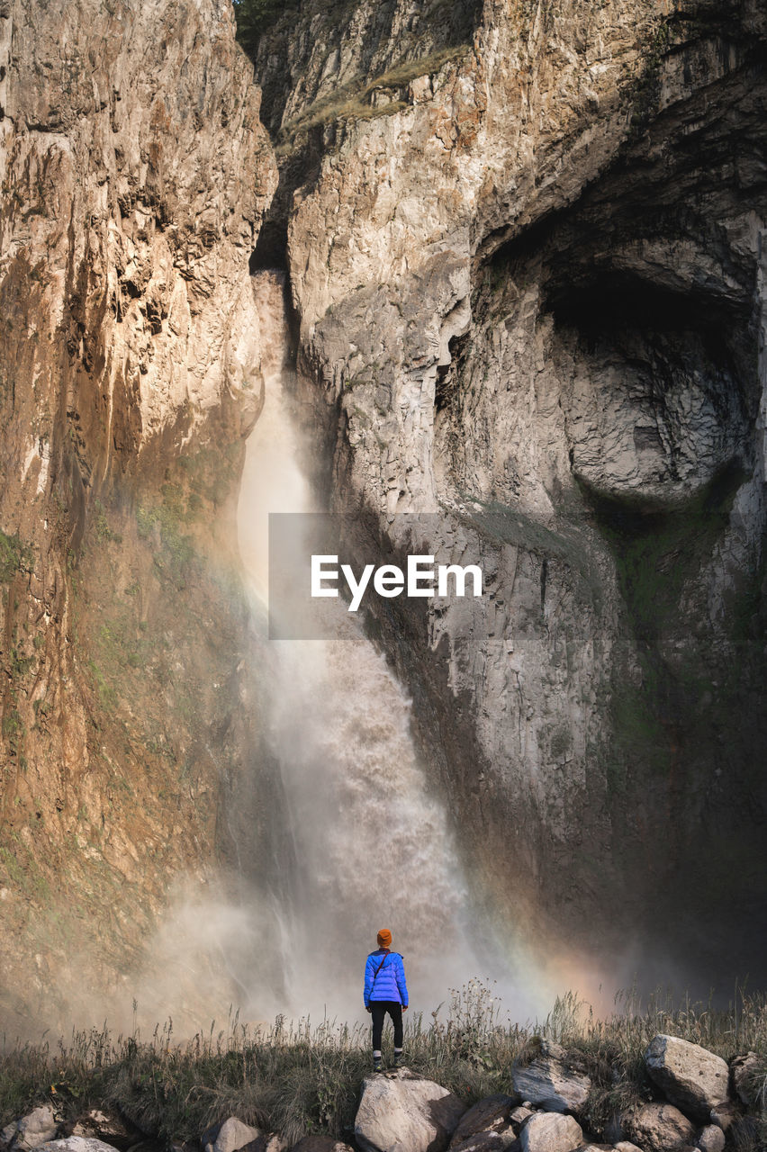 A woman in a warm jacket and hat stands with her back against the background of huge epic waterfall