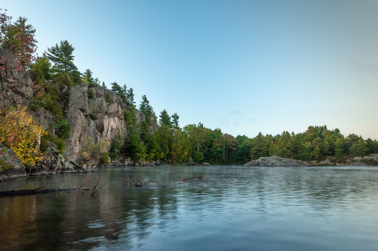 Calm lake surrounded by trees in front of mountains against clear blue sky