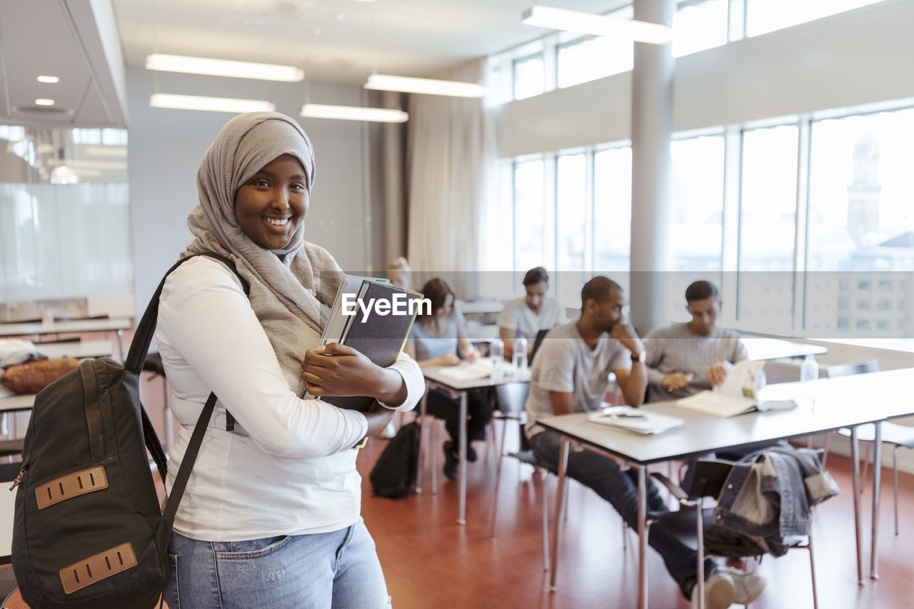 Portrait of smiling student in hijab standing with book against friends in classroom