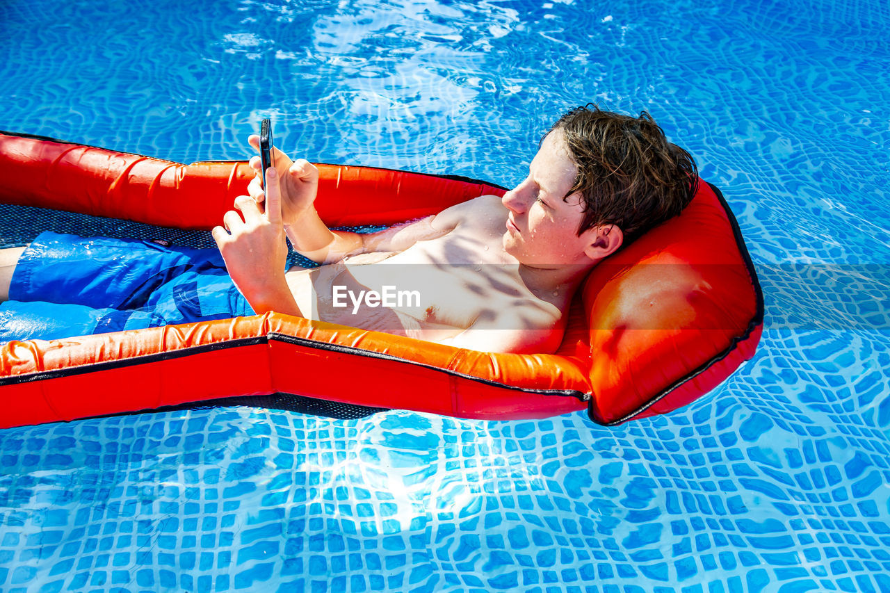 Boy floating on water in swimming pool using smartphone
