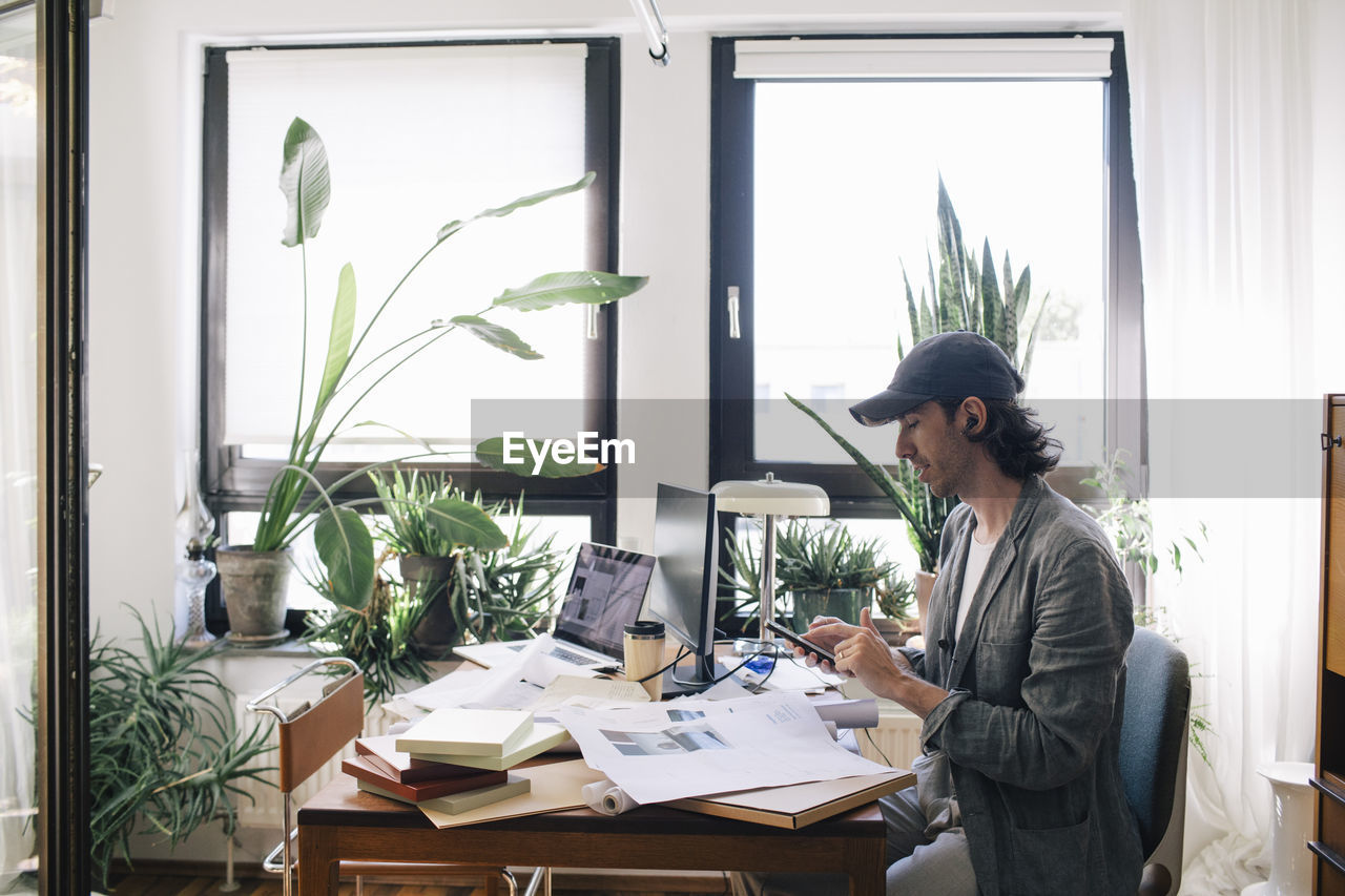 Side view of male architect using smart phone while sitting at desk in home office