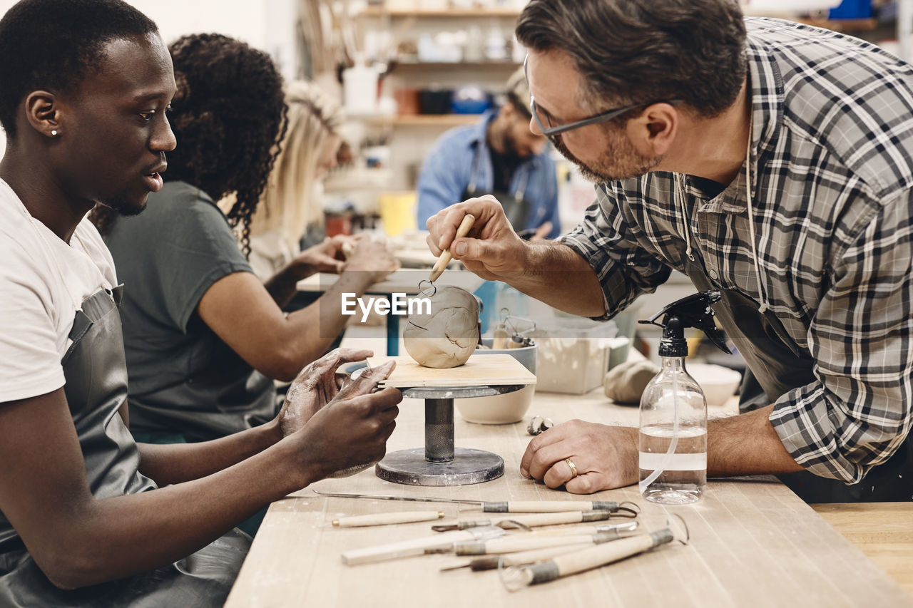 Mature instructor with work tool teaching student sitting at table in art class