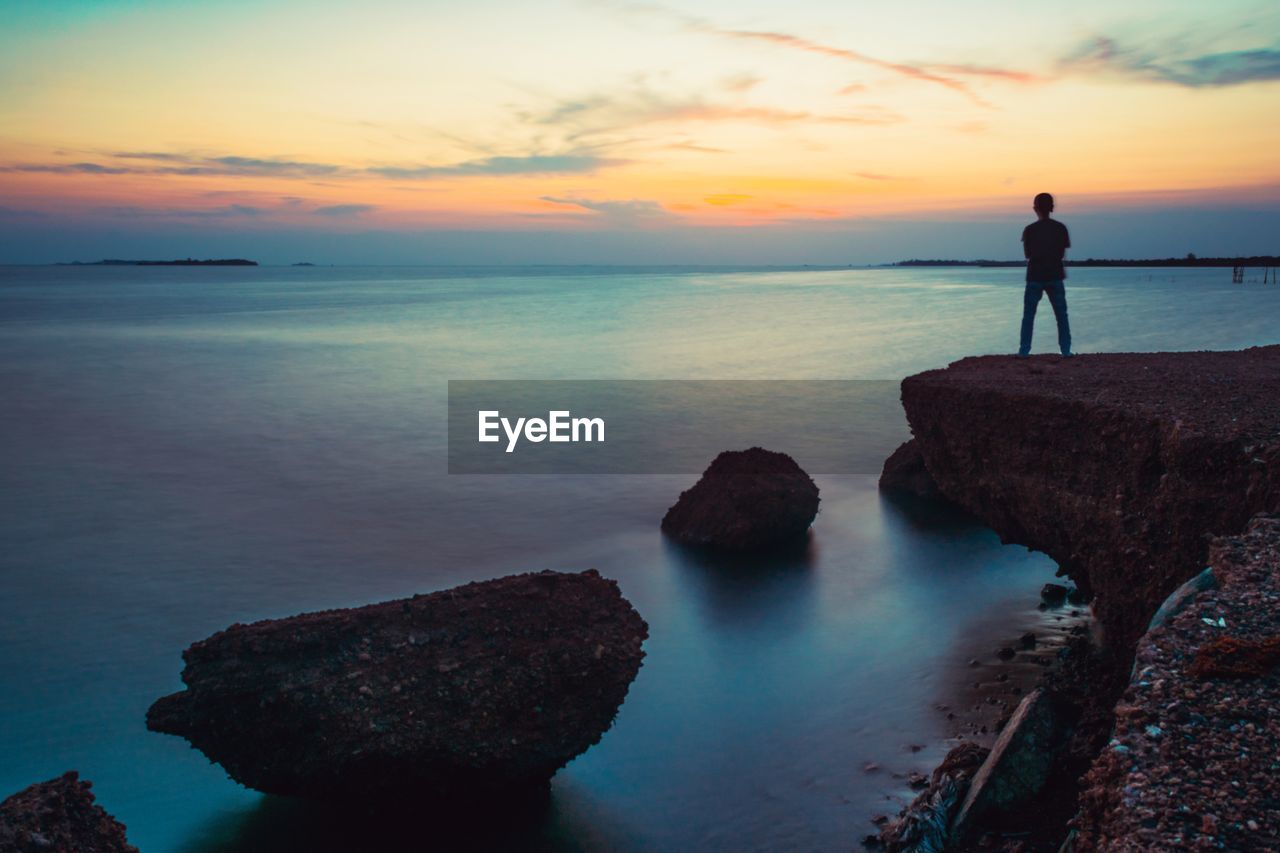 Rear view of silhouette man standing on cliff by sea against dramatic sky during sunset