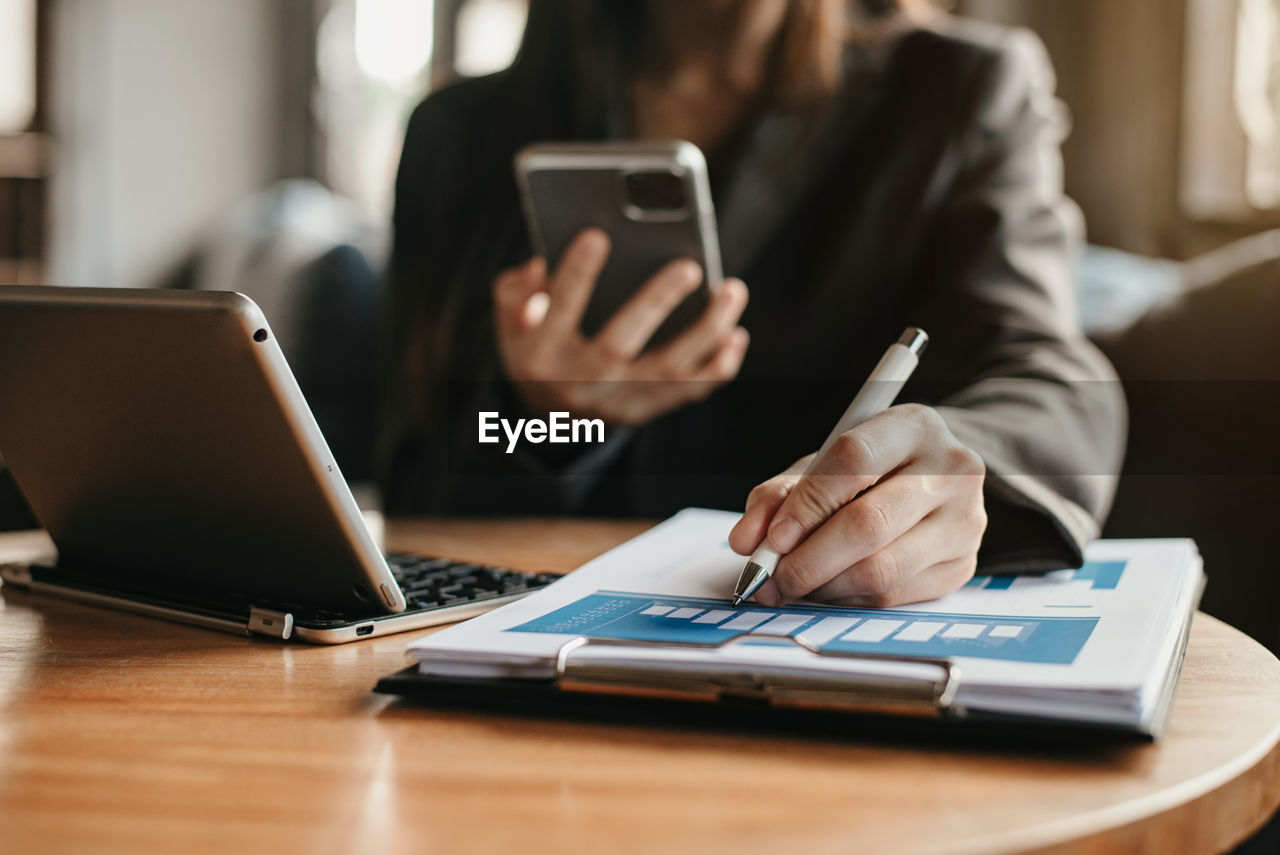 Midsection of woman holding smart phone writing on paper at desk