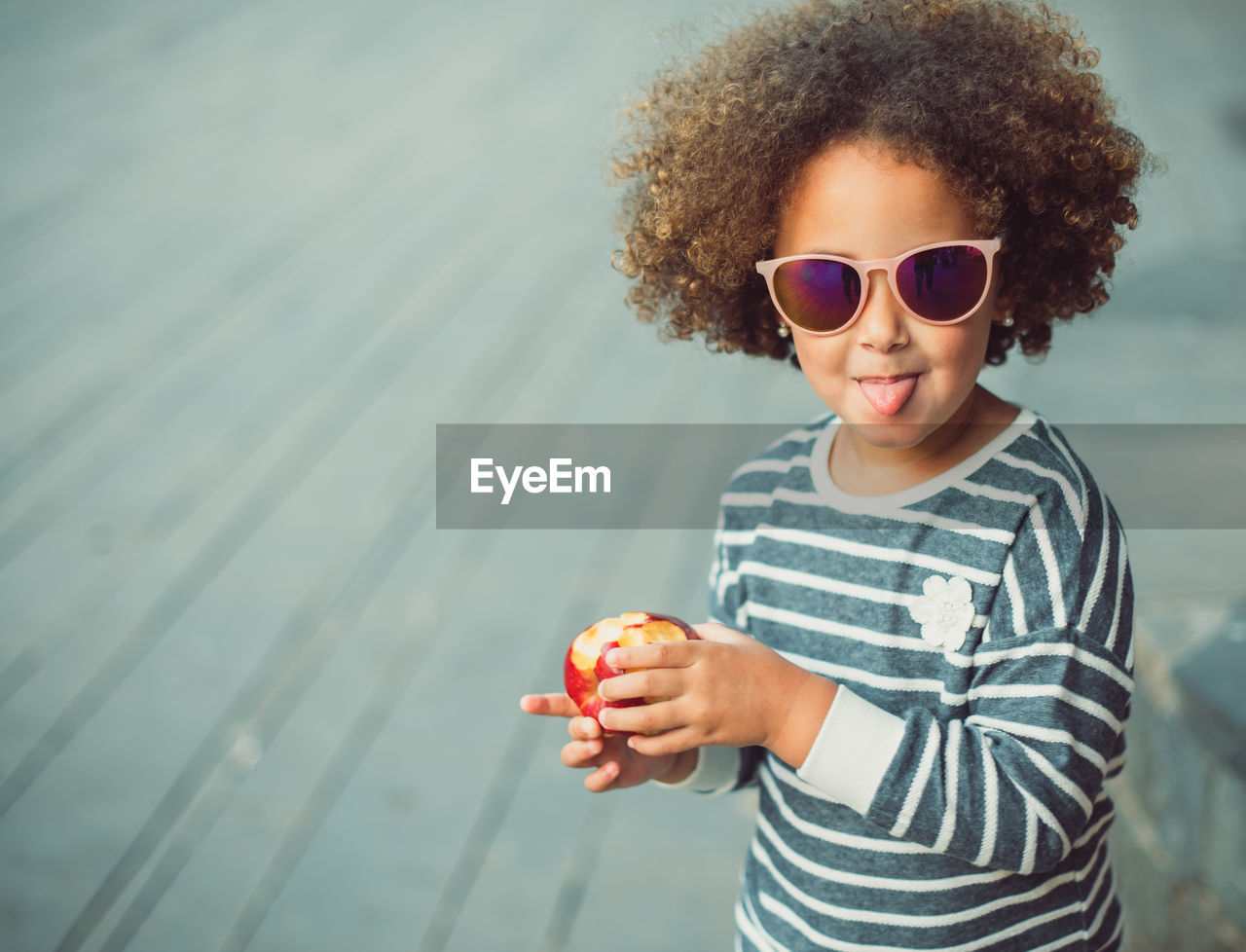 Cute little ethnic girl with afro hair wearing stylish striped shirt and sunglasses smiling and showing tongue while standing on city street and eating apple
