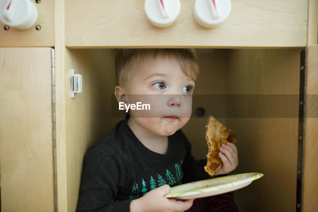 Toddler boy looks off into distance while eating pizza on paper plate