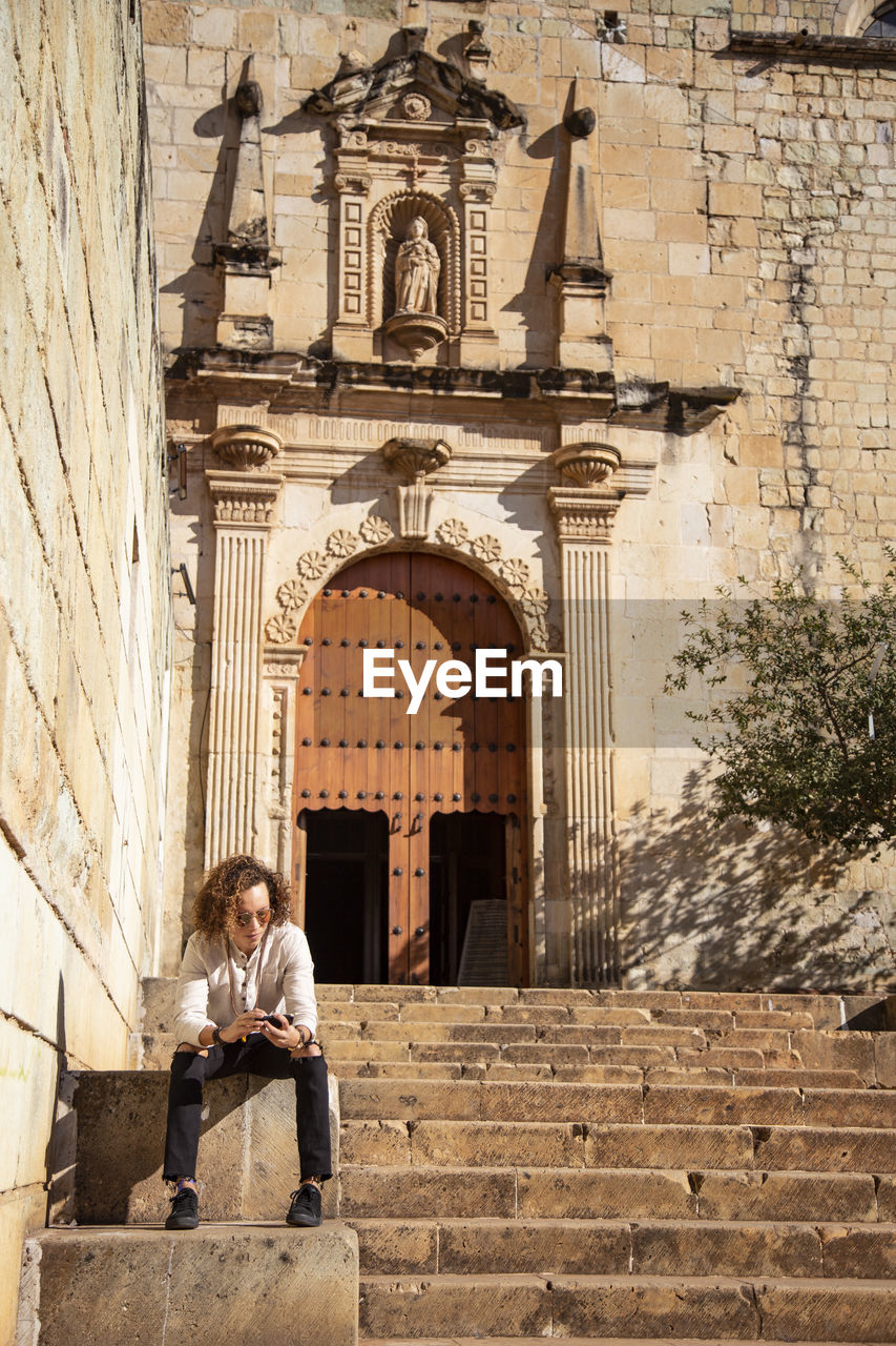 Man with iphone, sitted on staircase, outside baroque temple