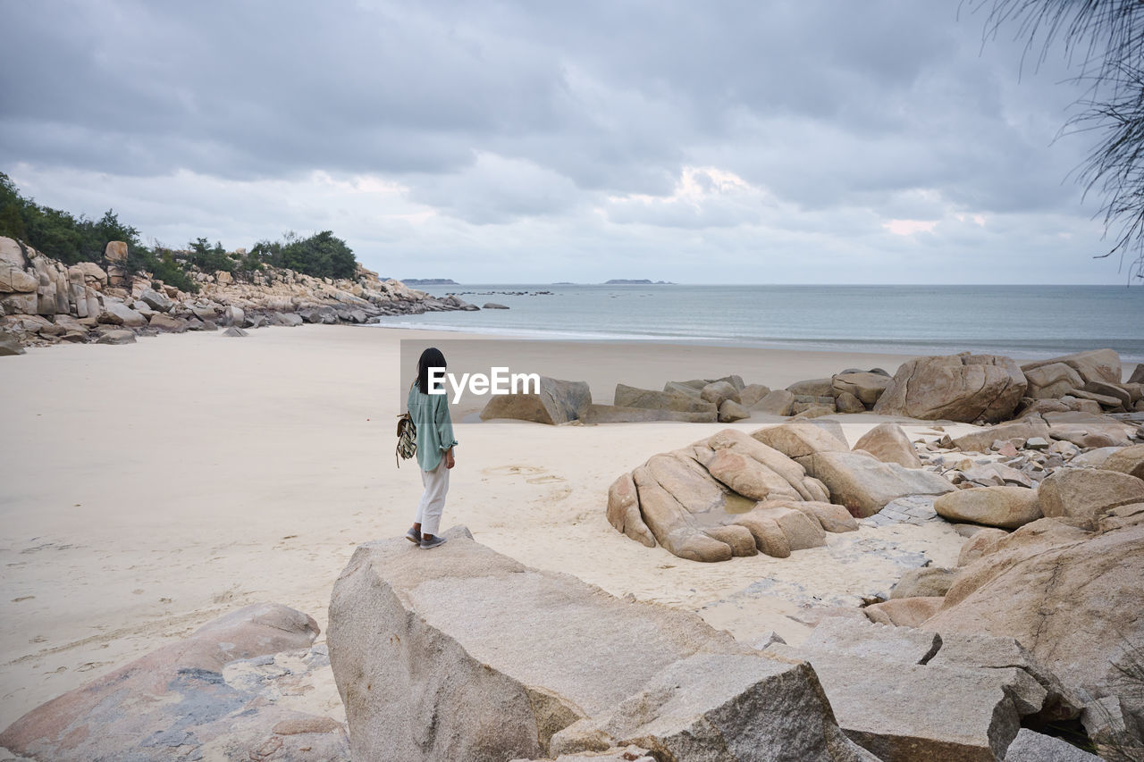 Side view of female standing on stone and admiring picturesque scenery of sea on cloudy day
