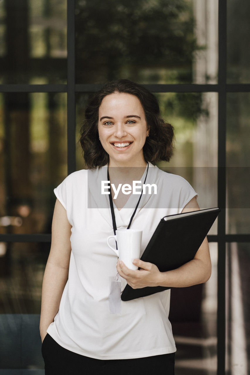 Portrait of happy young businesswoman standing with hand in pocket in front of glass wall at convention center