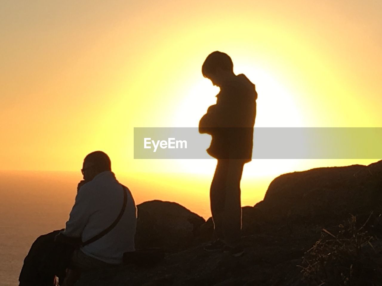 SILHOUETTE OF COUPLE SITTING ON LANDSCAPE AGAINST SUNSET