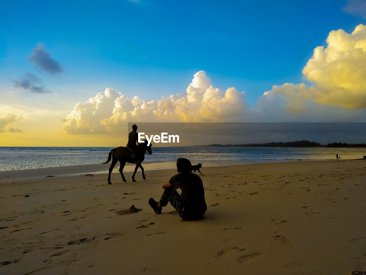 MAN SITTING ON BEACH AGAINST SKY DURING SUNSET