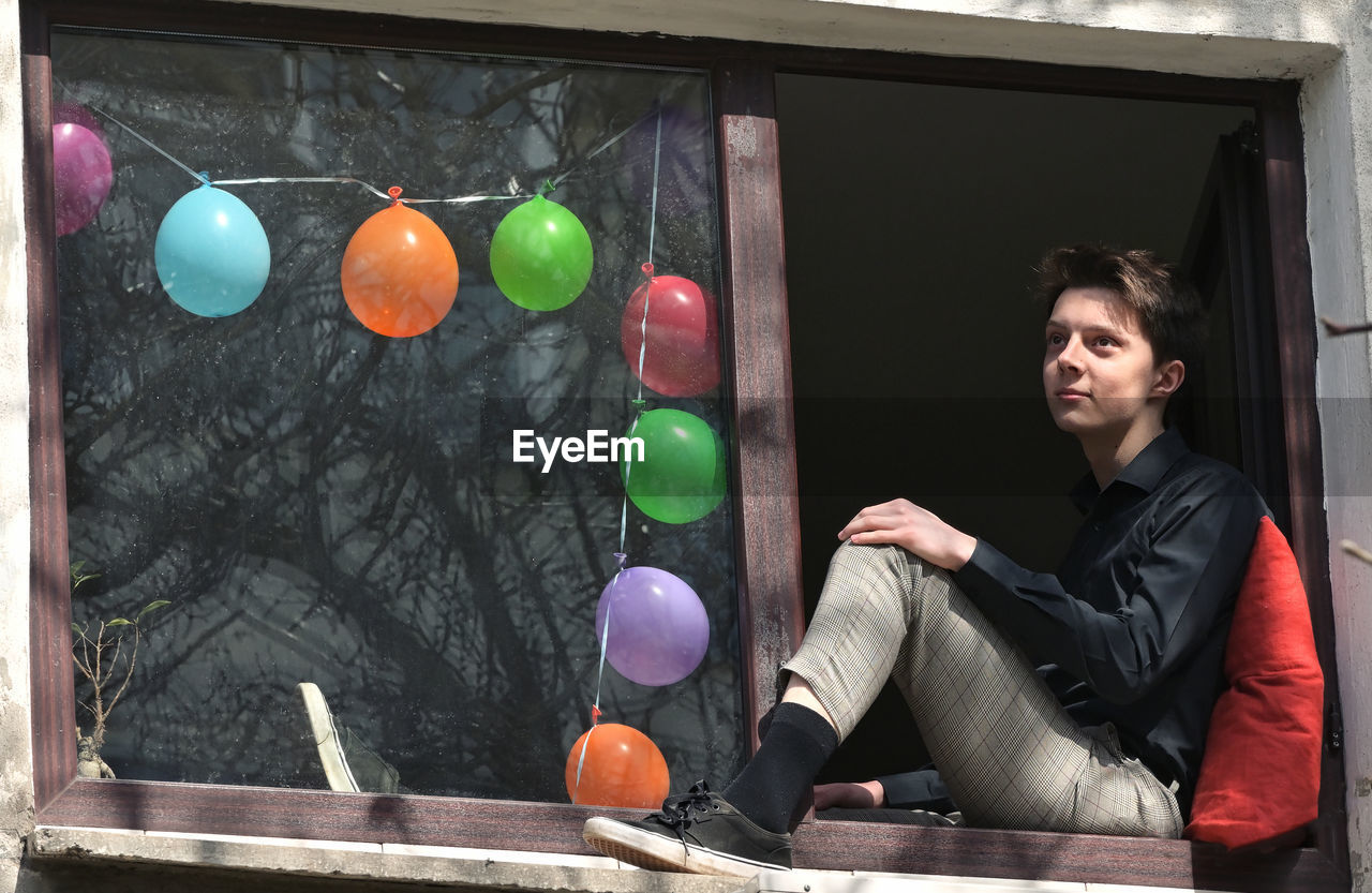 Thoughtful teenager sitting at window sill against multi colored balloons