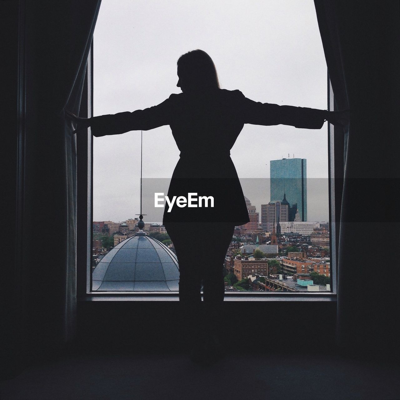 Rear view of silhouette woman standing near window in city against sky