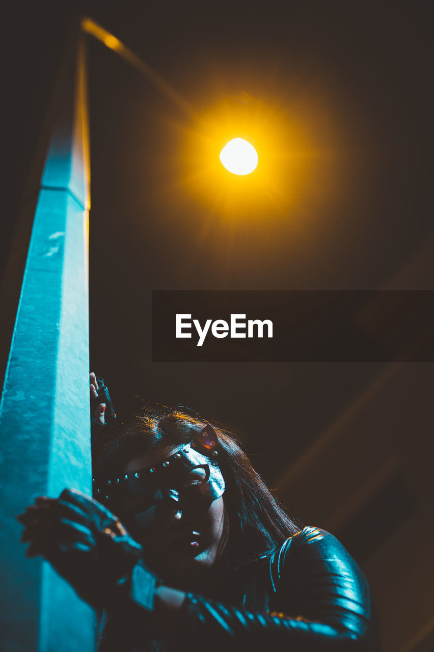 Low angle view of woman wearing mask while standing by illuminated street light at night