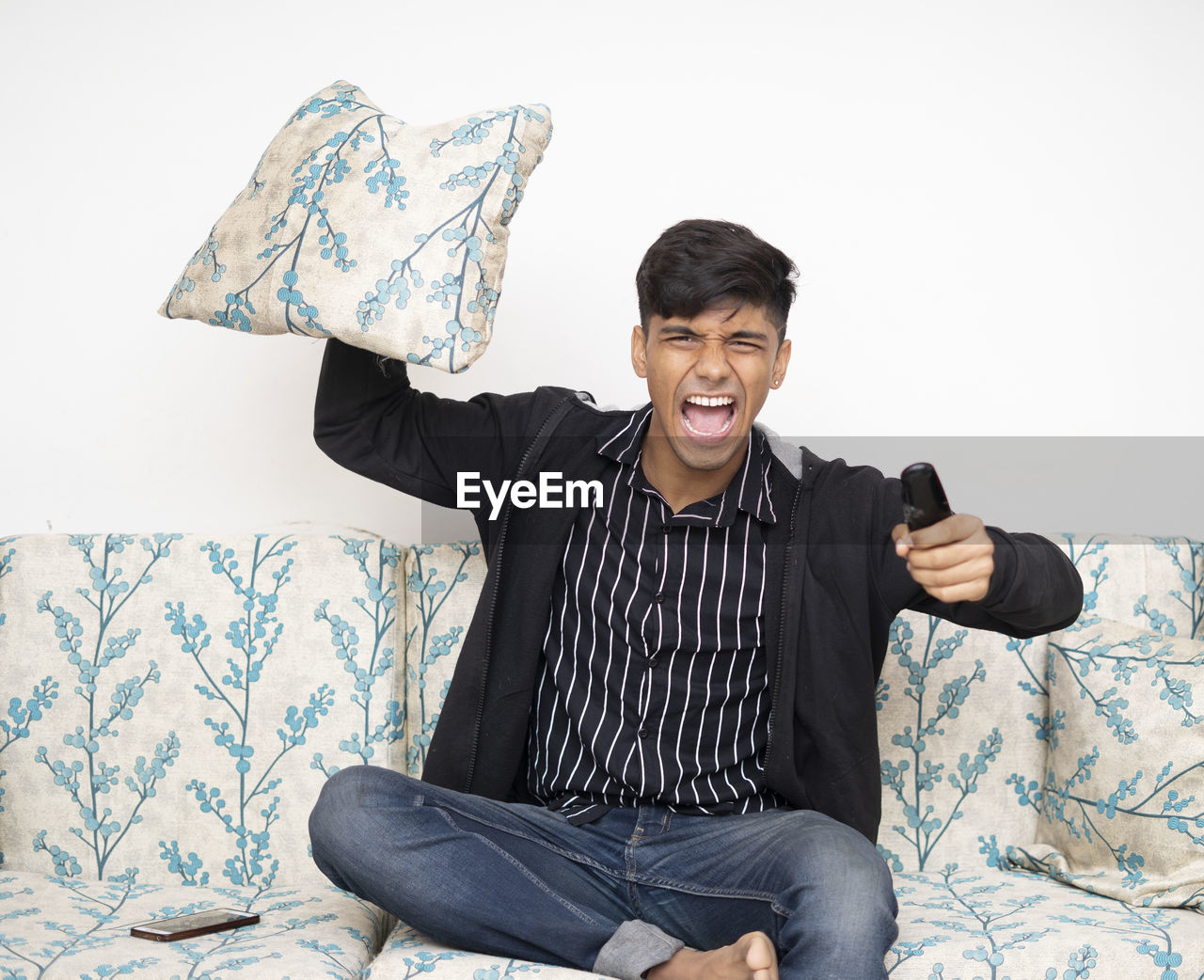 Asian man sitting on the sofa wearing black jacket holding remote while cheering for indian team.