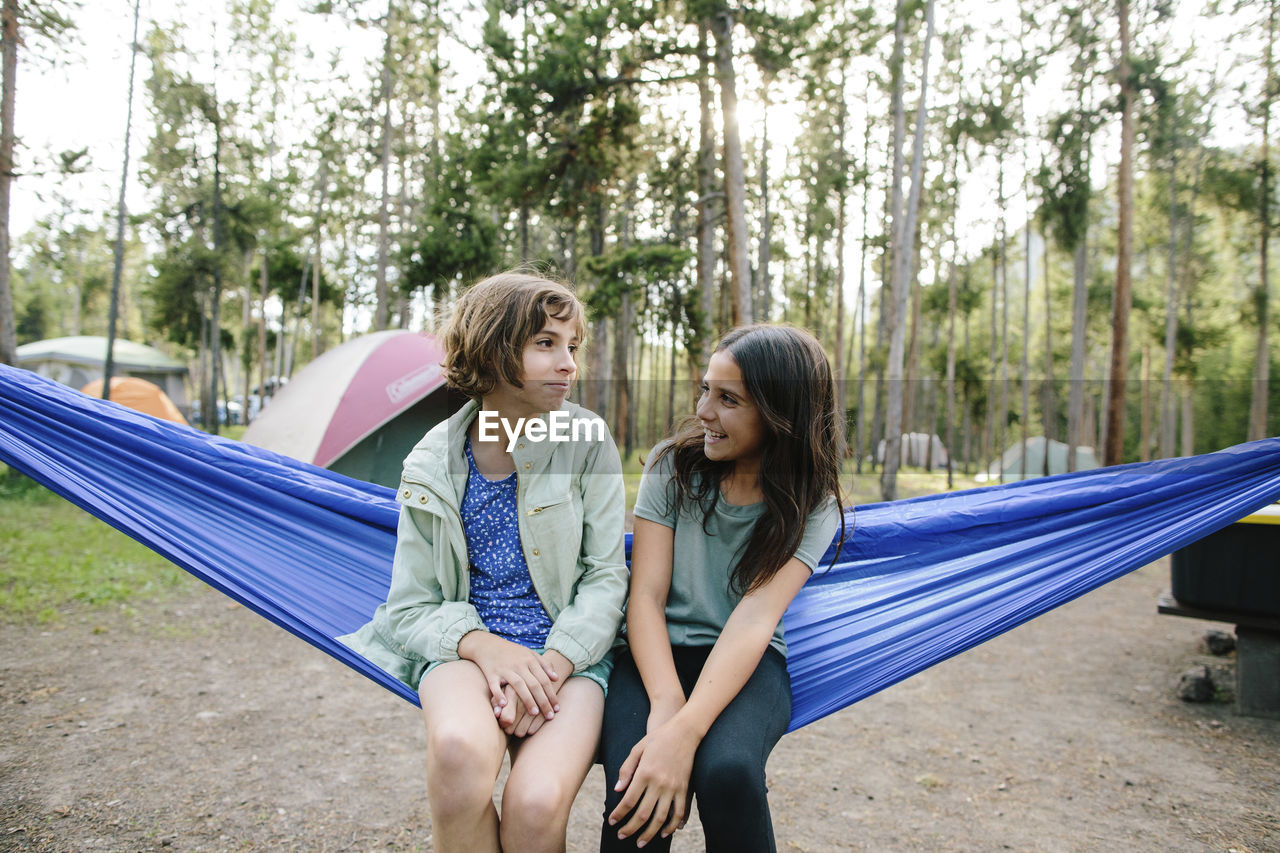 Sisters talking while sitting on hammock against trees in forest