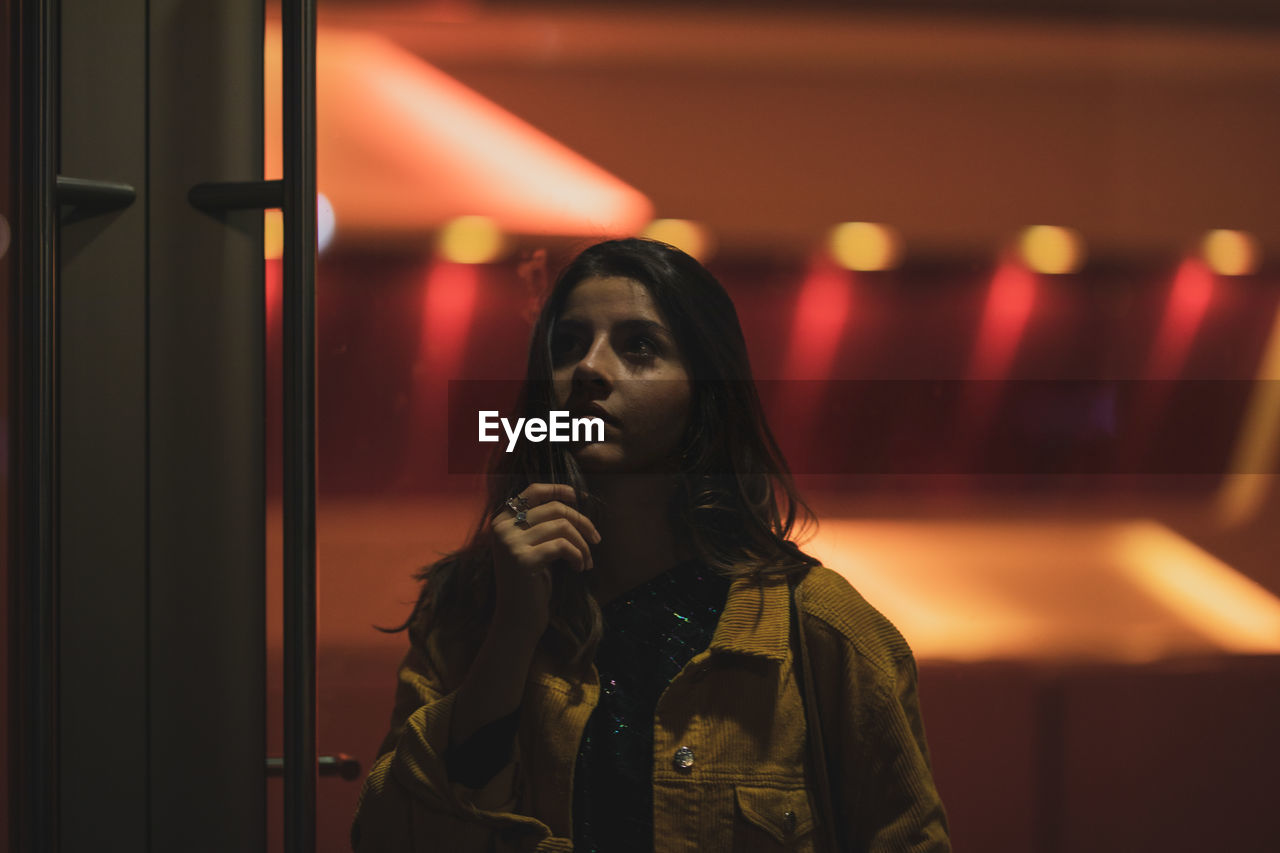 Young woman looking away while standing against illuminated lights at night