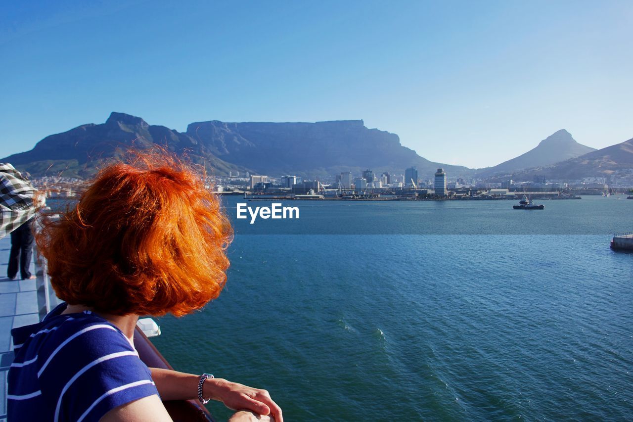 REAR VIEW OF WOMAN LOOKING AT SEA AGAINST MOUNTAIN RANGE