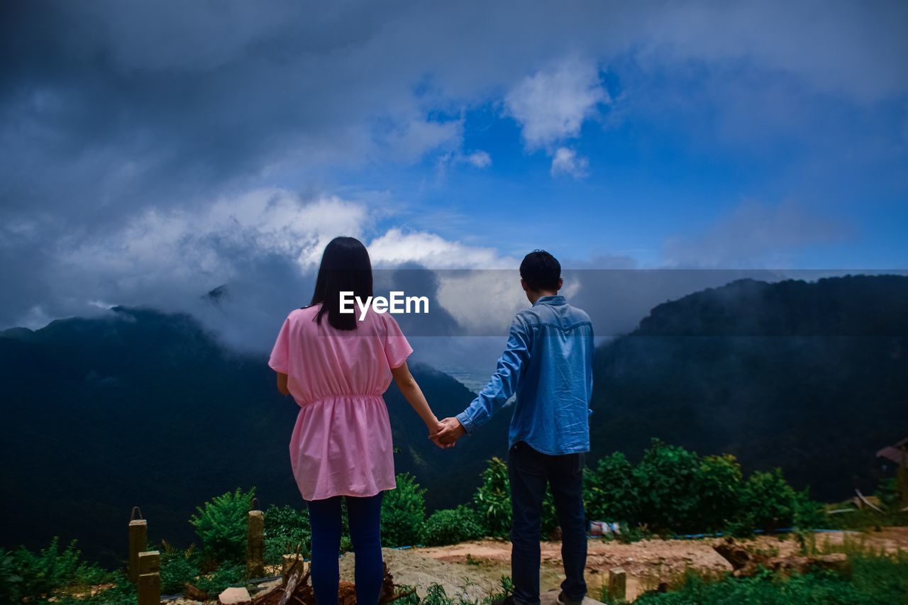 Rear view of couple holding hands standing on mountain against sky