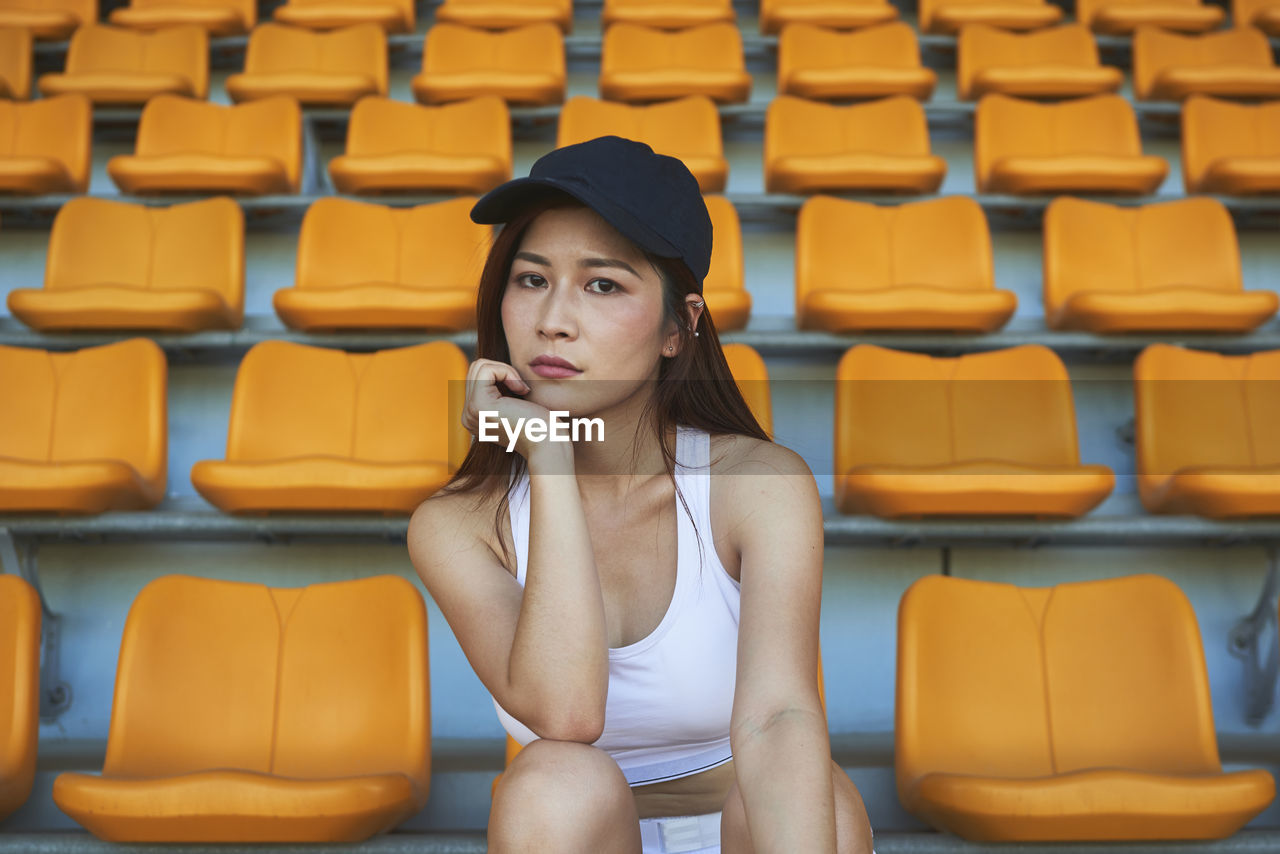 Confident asian athletic woman in sportswear sitting on stadium seat while resting after workout and looking at camera