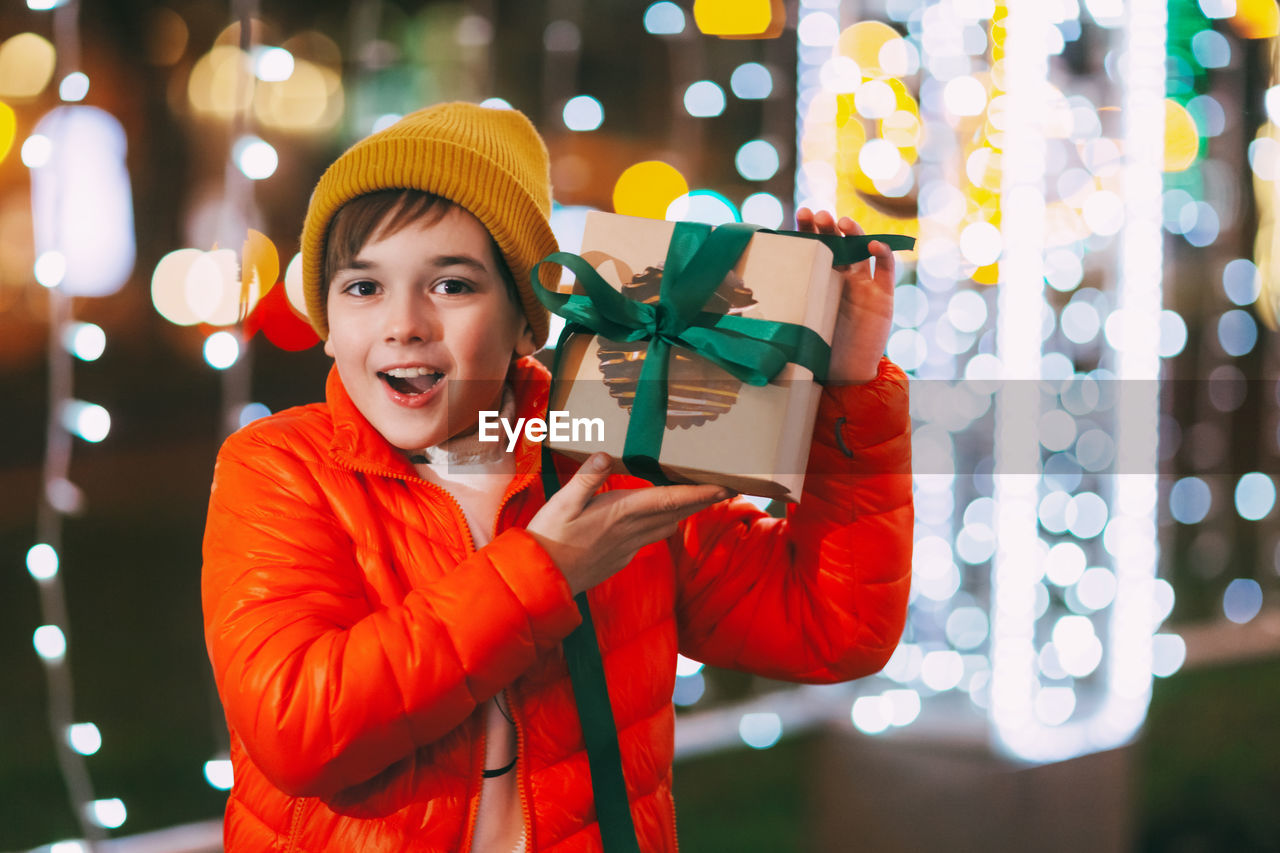 A happy boy in an orange jacket with a gift box in his hands at a christmas market 