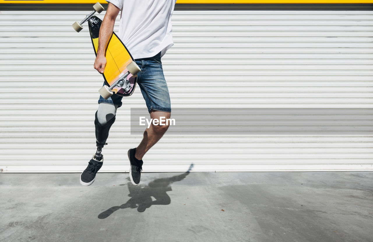Young man with leg prosthesis holding skateboard and jumping