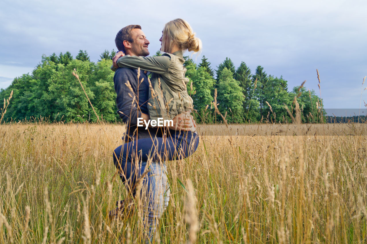 Couple kissing at grassy field against cloudy sky