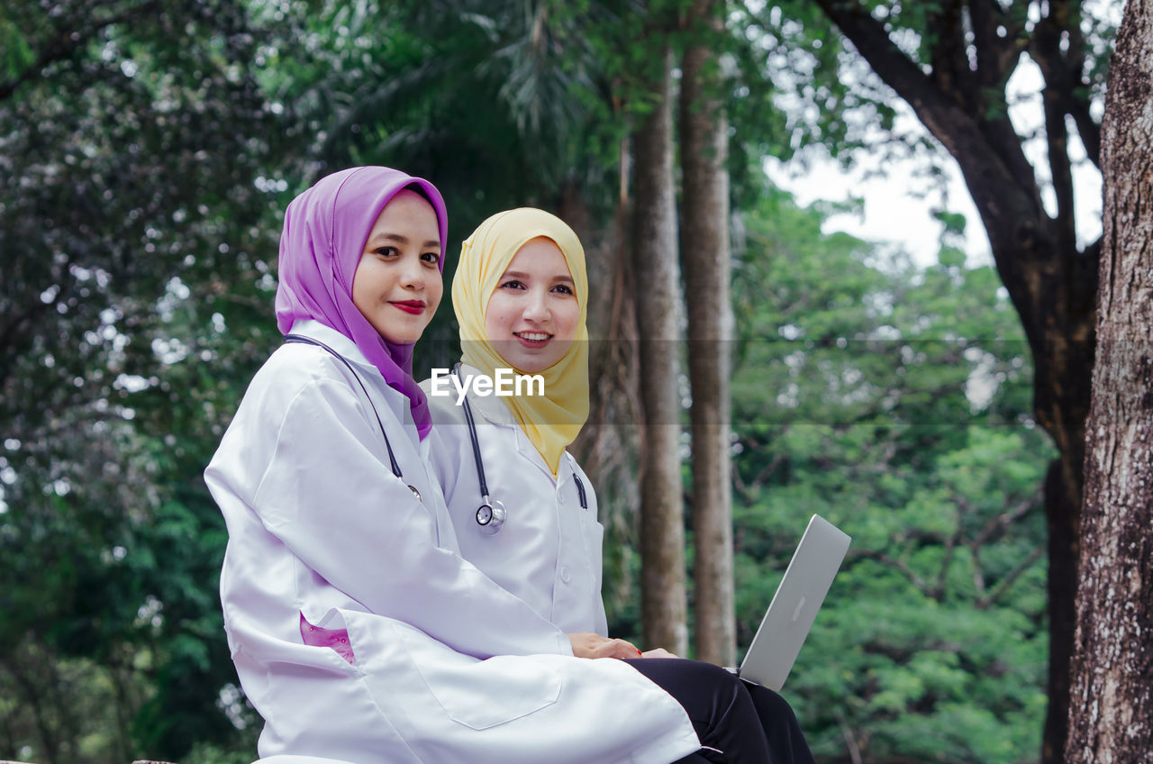 Portrait of female doctors using laptop against trees