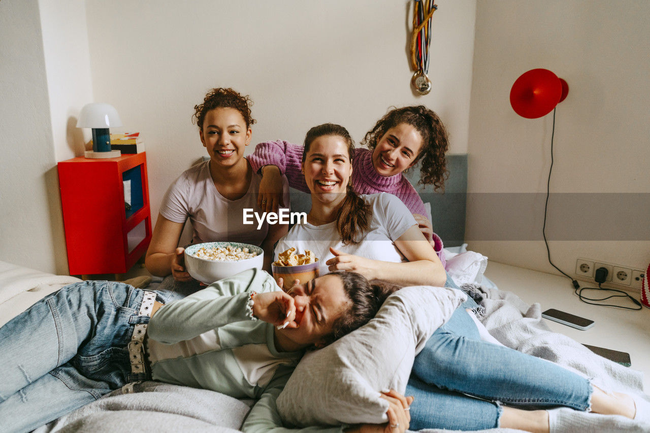 Happy female friends watching tv while enjoying snacks at home