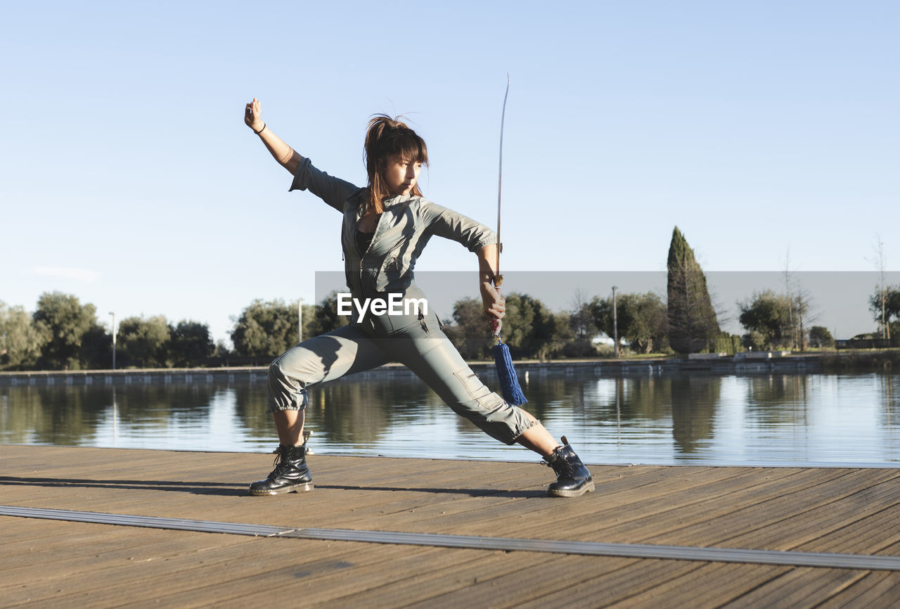 Confident female athlete with sword performing martial arts with dedication by lake on sunny day