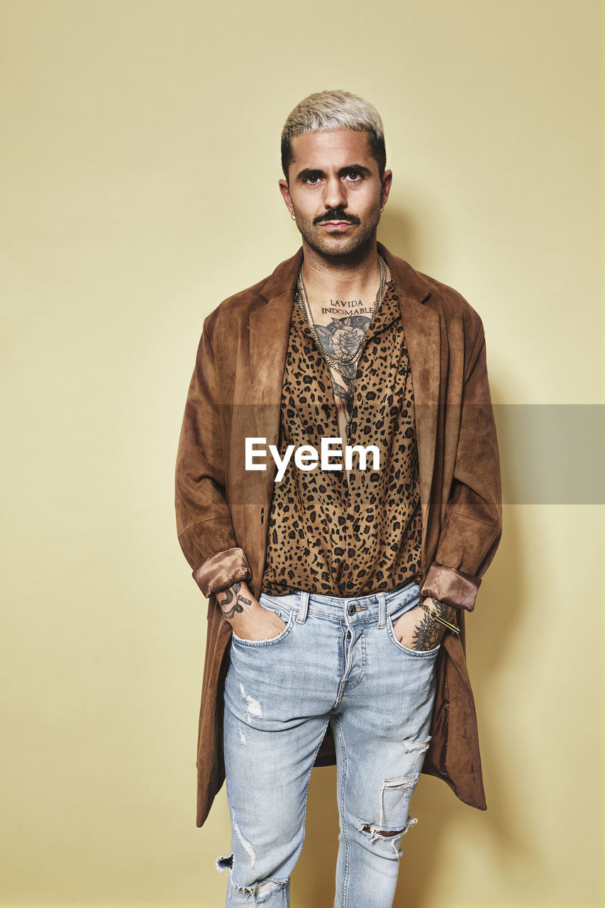 Fashionable male model with tattoos wearing trendy coat over leopard shirt and jeans standing against beige background and looking at camera