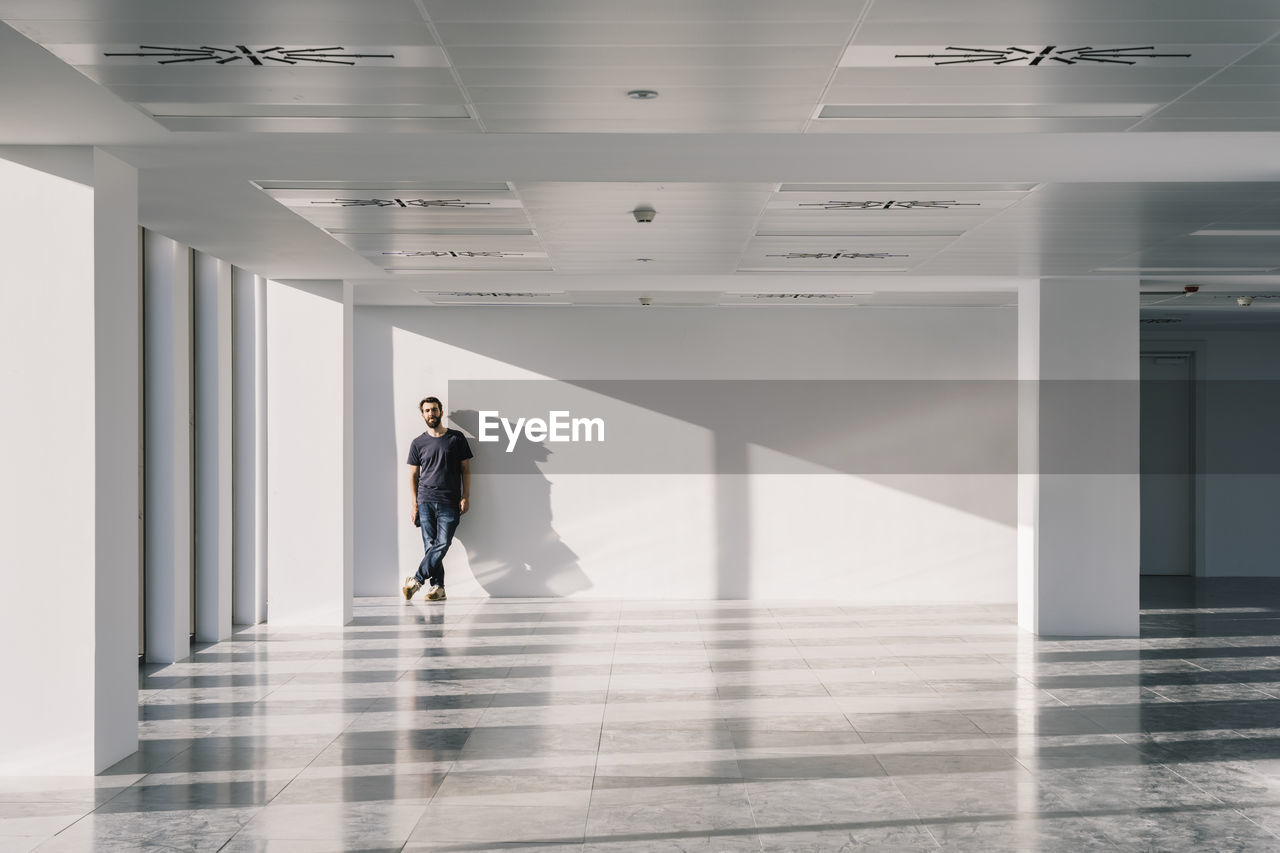 Male standing near window in empty spacious office hallway with shadows and sunlight on white walls and looking at camera