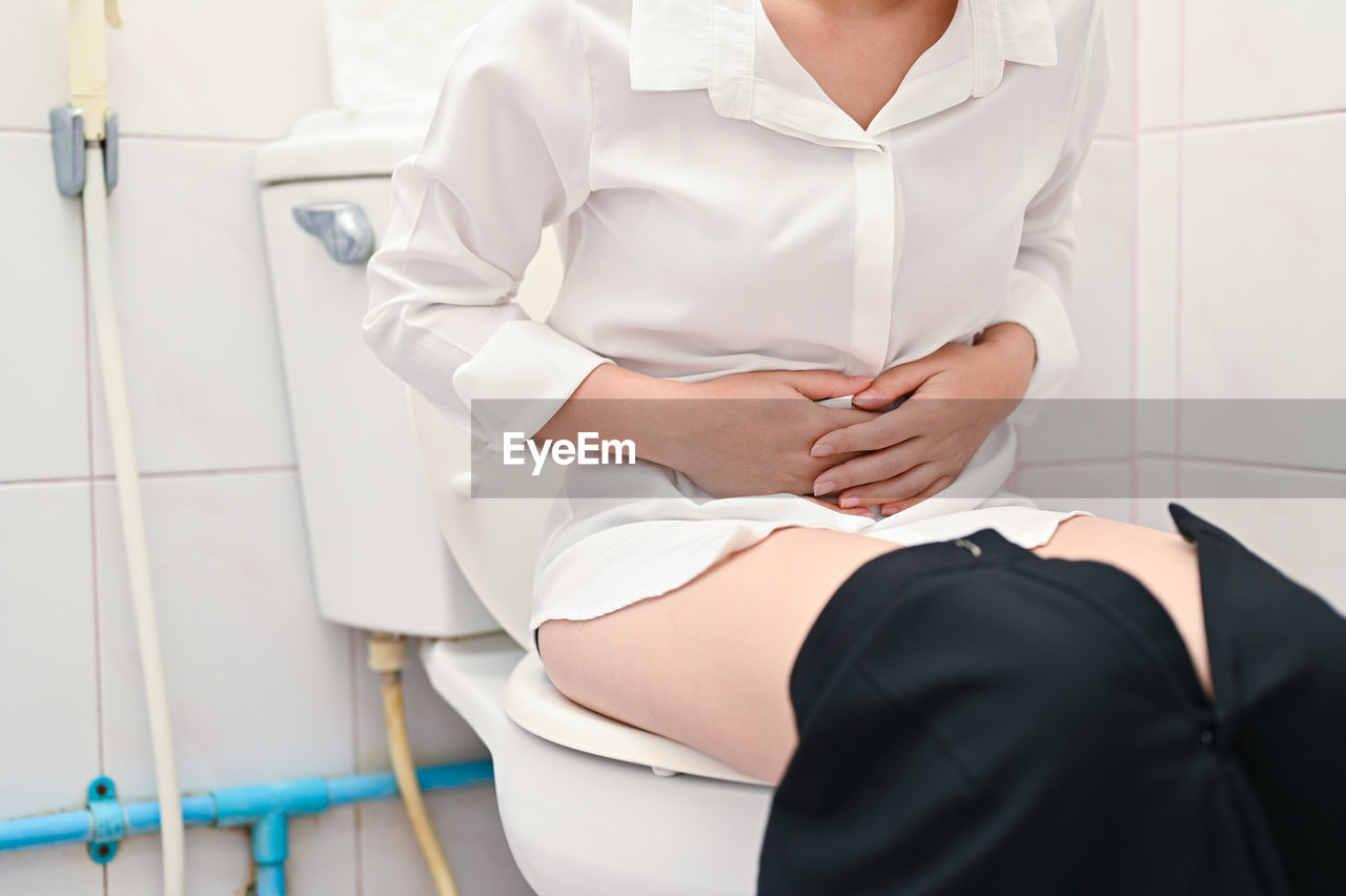 Midsection of woman sitting in bathroom while holding stomach at home