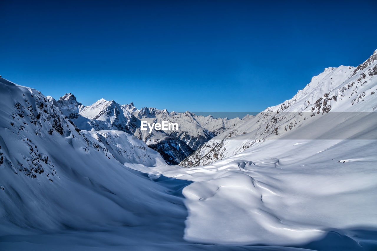 snow, cold temperature, winter, mountain, scenics - nature, landscape, environment, beauty in nature, mountain range, sky, nature, blue, snowcapped mountain, ice, frozen, travel destinations, tranquil scene, tranquility, no people, clear sky, travel, screenshot, piste, land, non-urban scene, extreme terrain, outdoors, glacier, sunny, mountain peak, idyllic, winter sports, sunlight, white, cloud, ski touring, copy space, day, panoramic