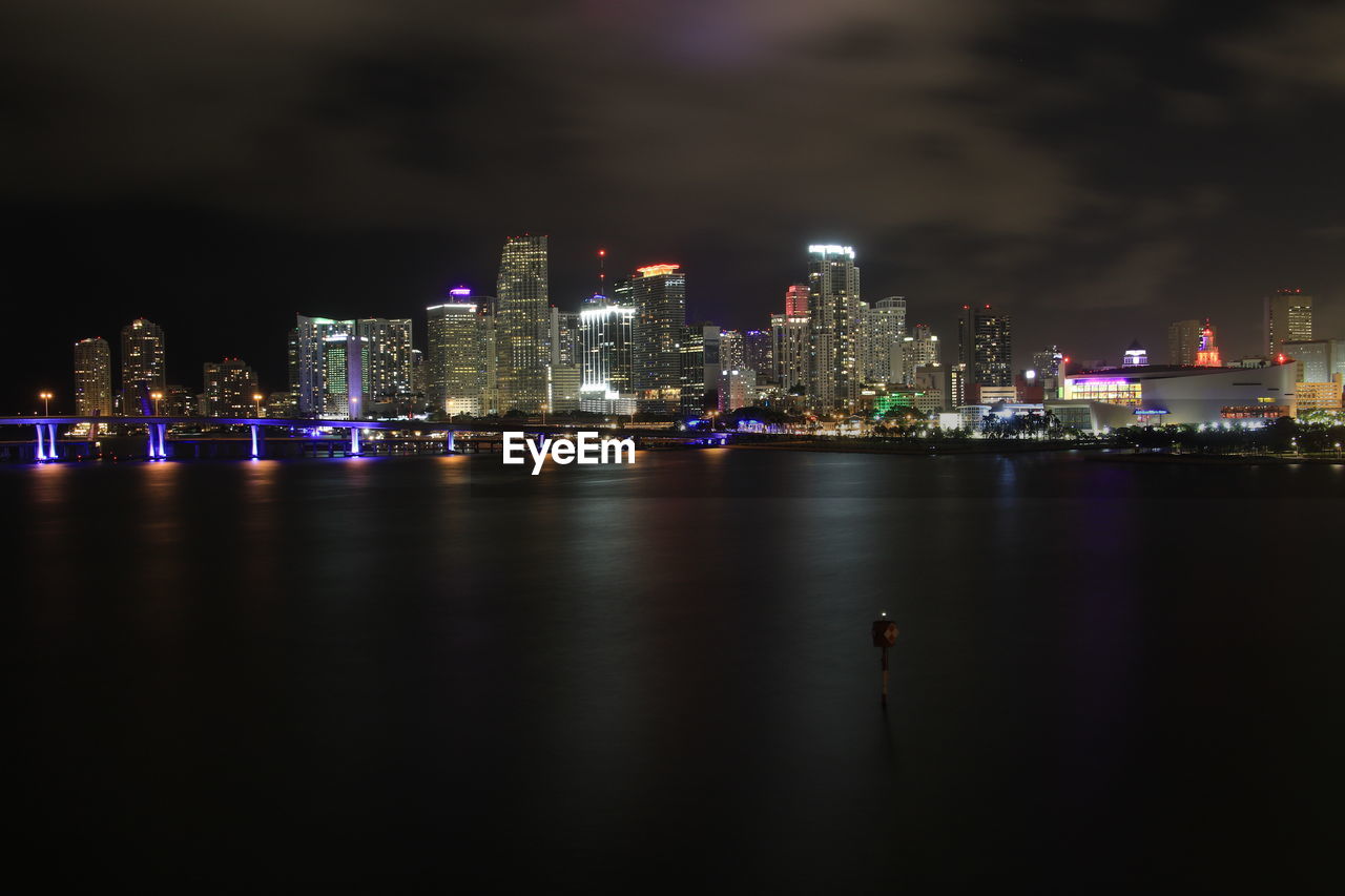 ILLUMINATED BUILDINGS BY RIVER AGAINST SKY