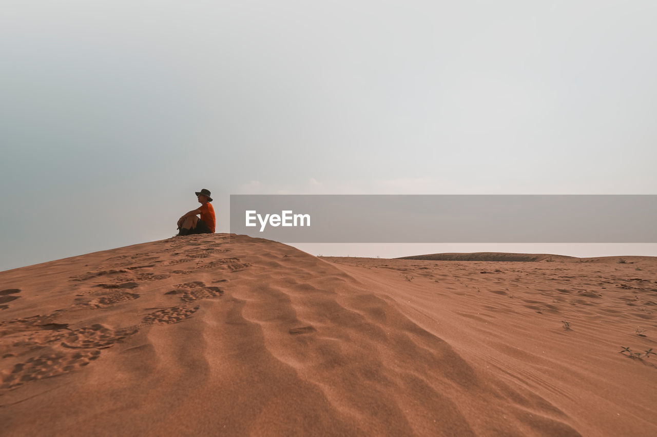 Side view of mature man sitting on sand dune at desert against sky