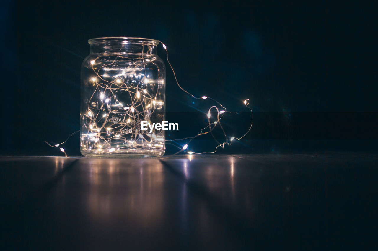 CLOSE-UP OF GLASS JAR ON TABLE AGAINST ILLUMINATED BACKGROUND
