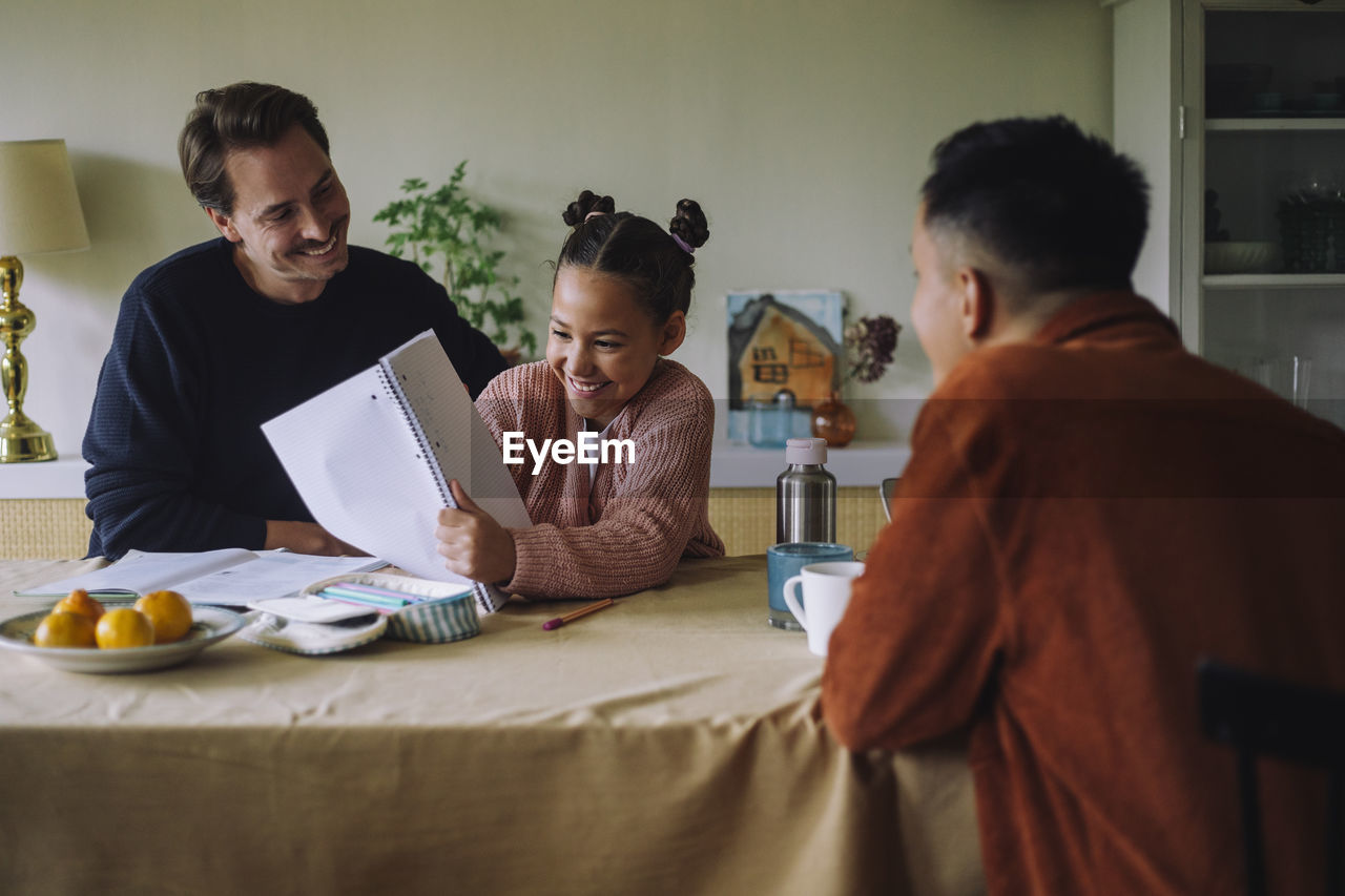 Happy daughter showing notebook to gay father at dining table in home