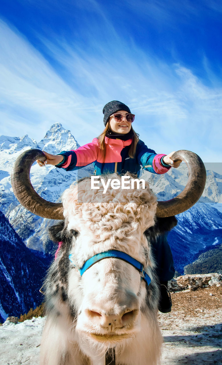 Smiling woman wearing knit hat sitting on highland cattle against sky