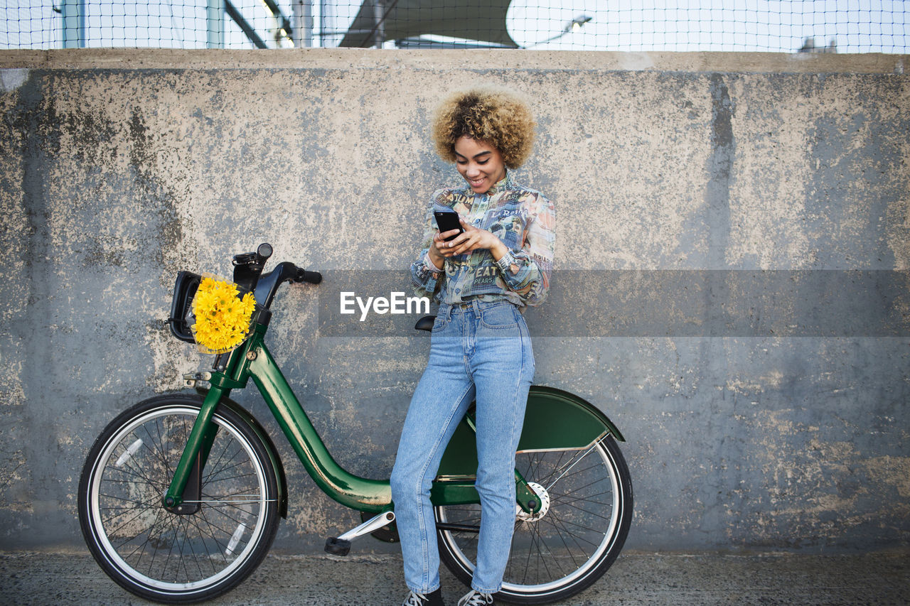 Woman using phone while standing by bicycle against wall