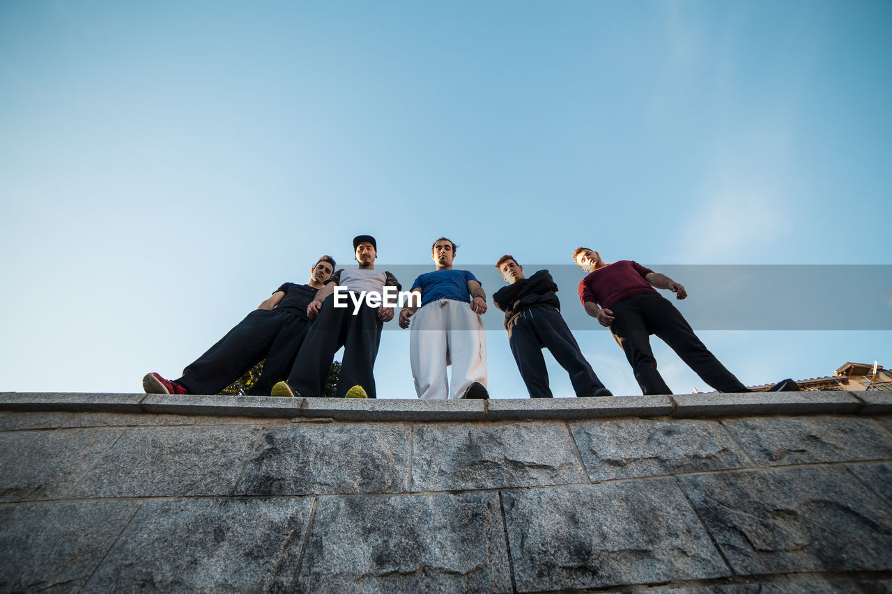 Low angle view of male friends standing on retaining wall against sky