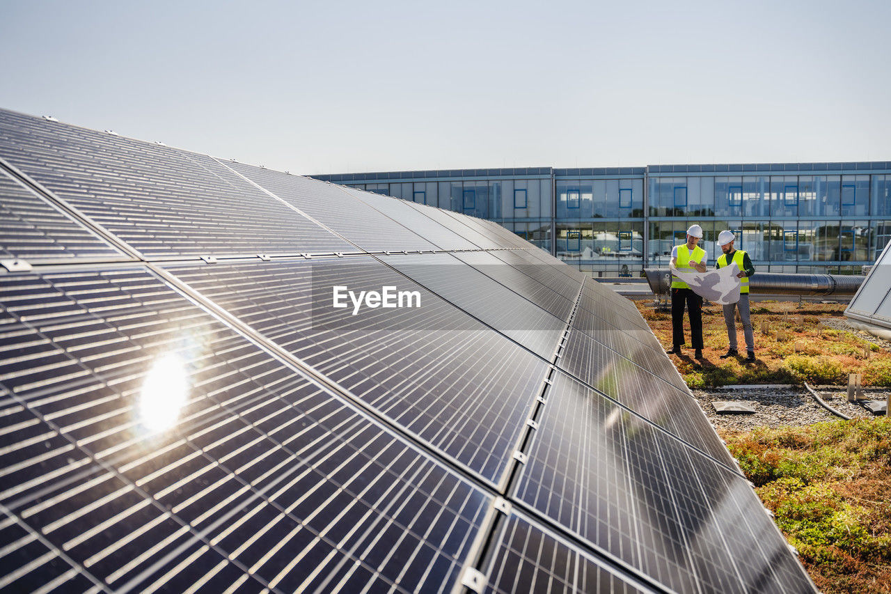 Two technicians analyzing blueprints on the rooftop of a corporate building equipped with solar panels