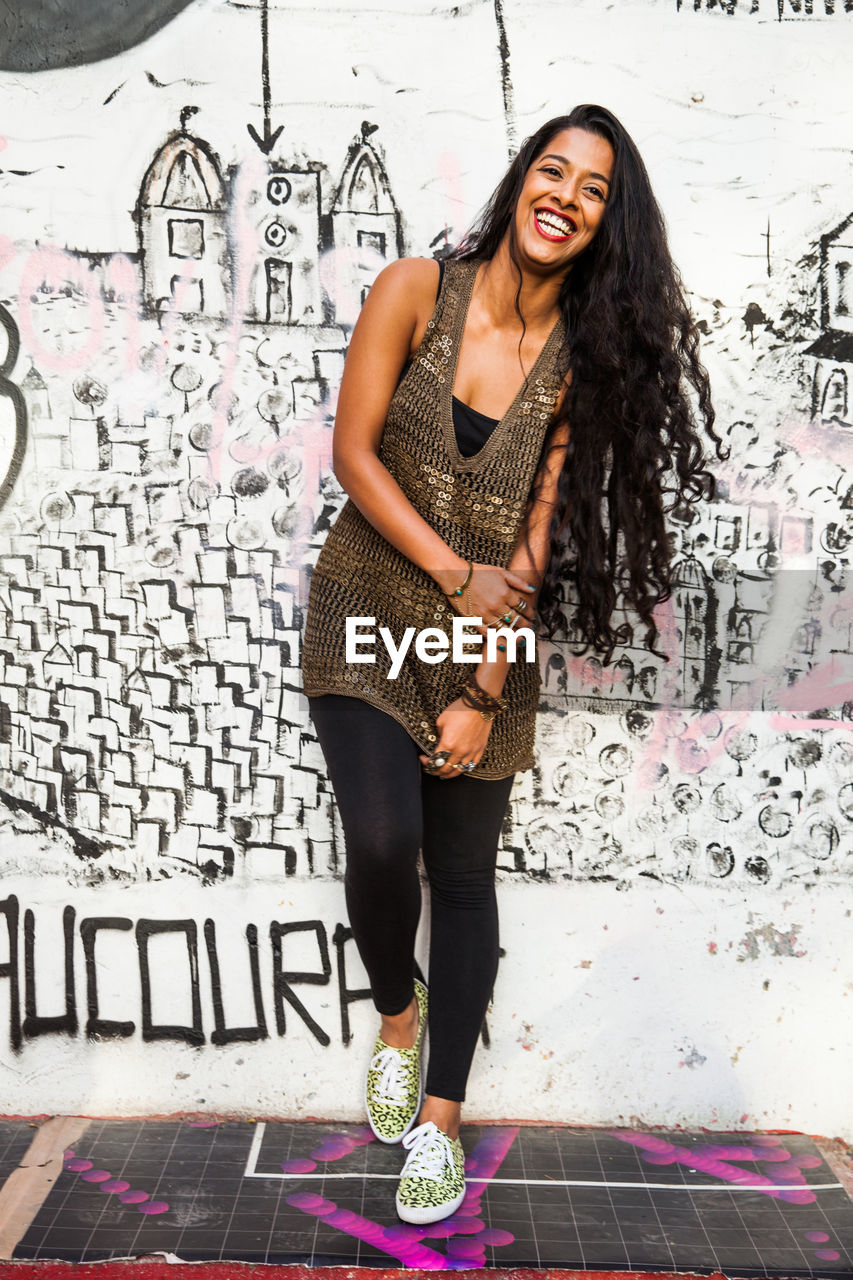 PORTRAIT OF A SMILING YOUNG WOMAN AGAINST WALL