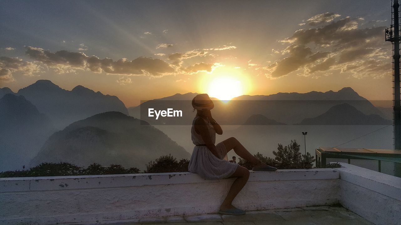 Young woman sitting on building terrace against sky during sunset