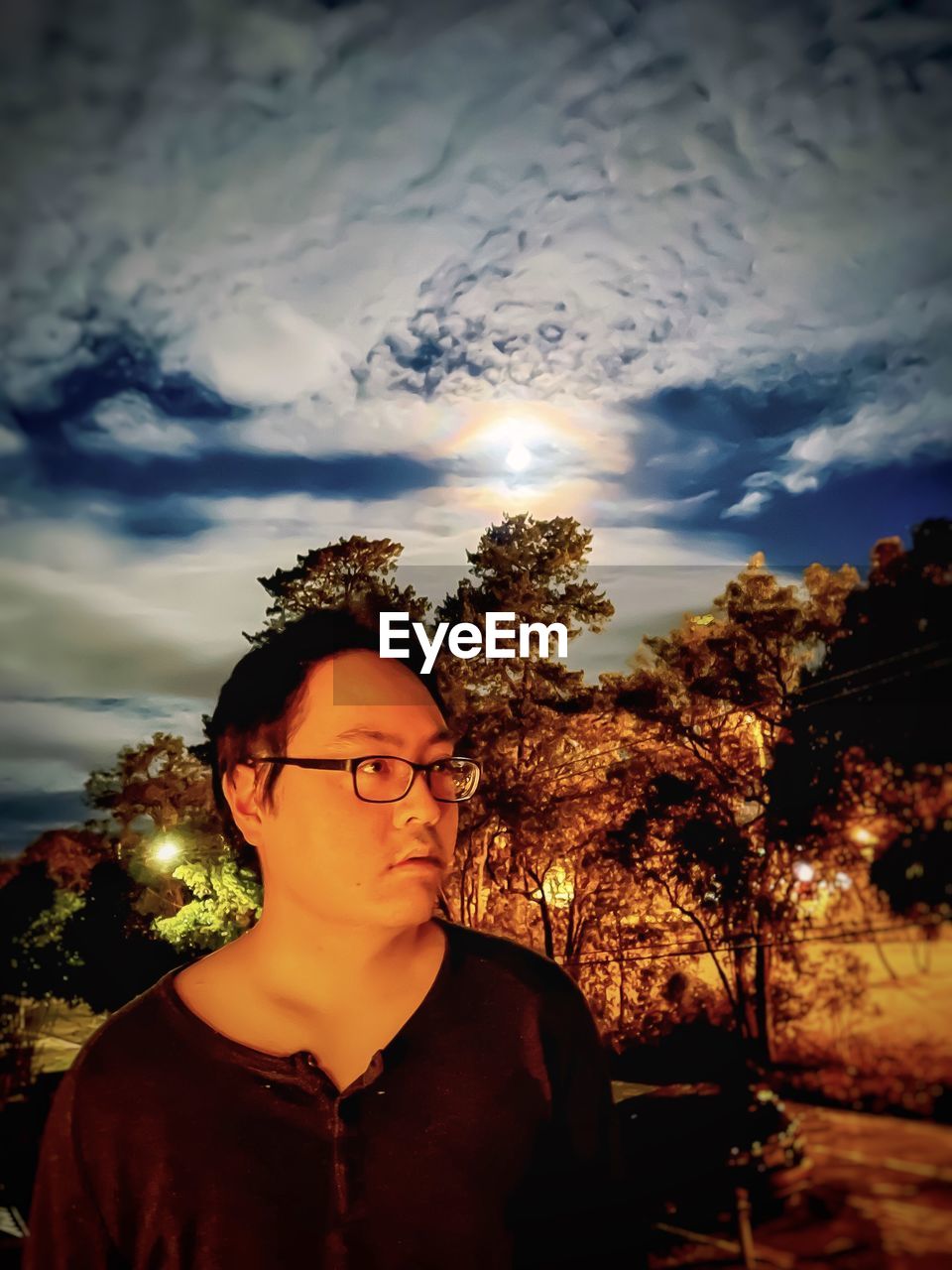 Portrait of young man standing against cloudy sky, rainbow colored lunar halo and trees at night.