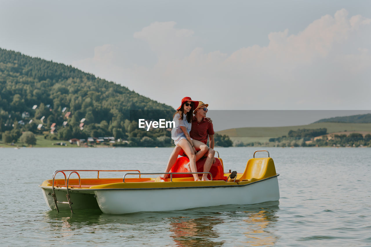 Cheerful man and woman in paddleboat on lake against sky