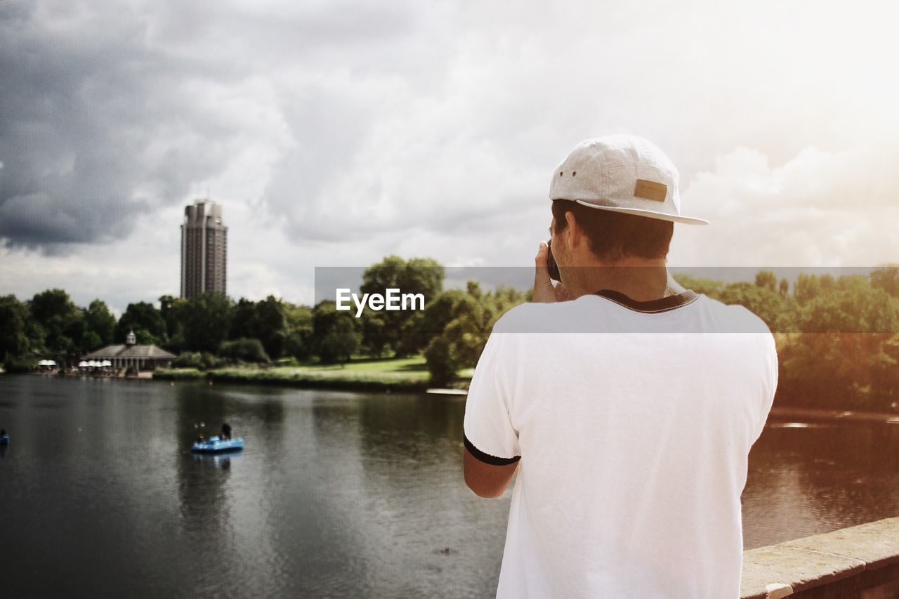 Rear view of man photographing building and river against cloudy sky