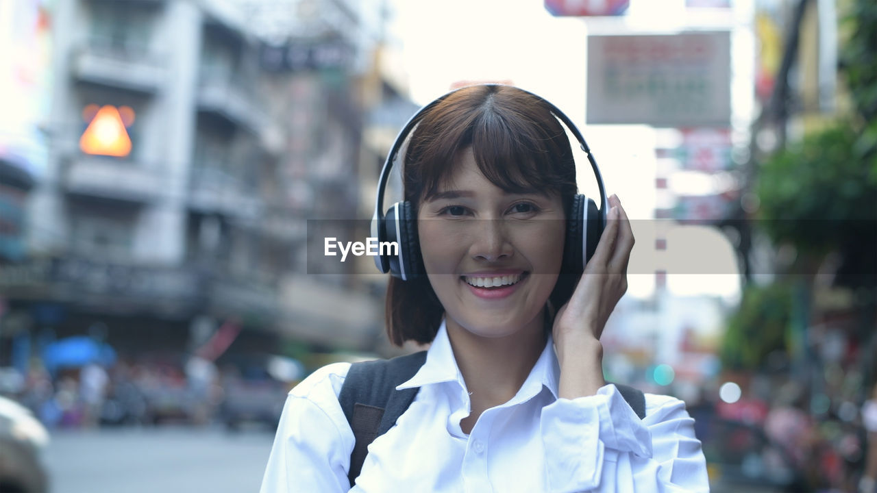 Portrait of smiling woman listening music through headphones while standing in city