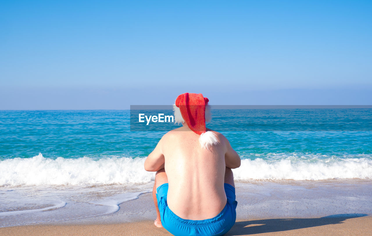 A man in a santa claus hat sits on the seashore, rear view. christmas santa claus relaxing